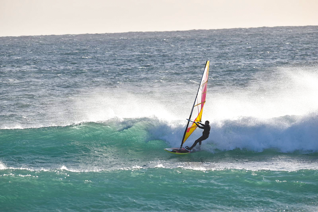 A windsurfer rides across a breaking wave in choppy ocean waters under an overcast, gray sky. The windsurfer is viewed from behind, gripping the boom of a vibrant sail featuring bright yellow, orange, and pink panels.